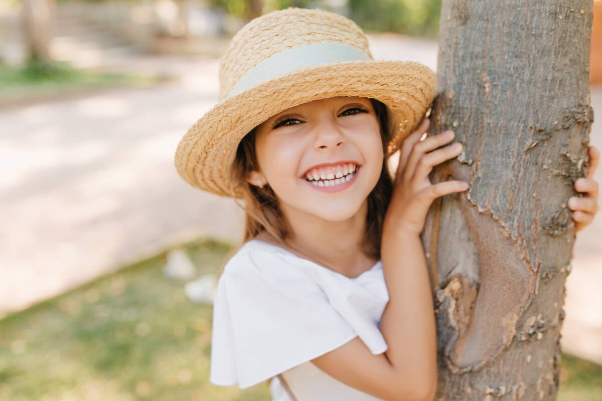 laughing little girl with lightly tanned skin posing park touching tree outdoor close up portrait cheerful dark haired kid vintage hat with ribbon having fun garden